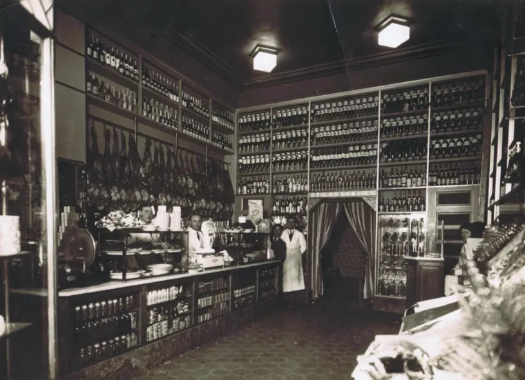 El interior en blanco y negro: jamones colgados sobre el mostrador, las estanterías llenas de conservas y botellas, y el personal detrás de la barra.